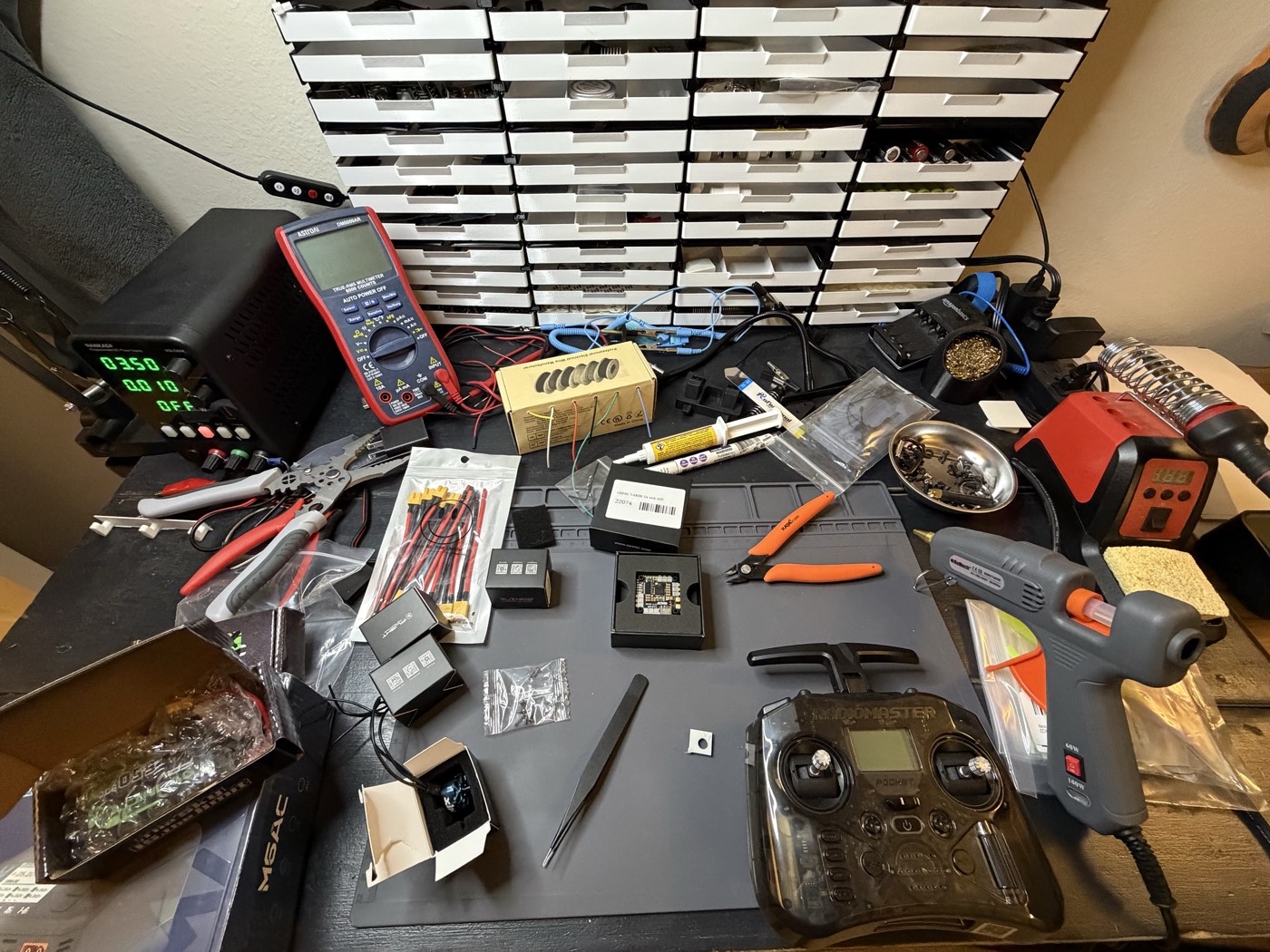 A workbench laid out with drone-building gear — a bench power supply, multimeter, soldering iron station, hot glue gun, pliers, a RadioMaster radio controller, a flight controller board, ESCs and battery leads — in front of labeled parts drawers.